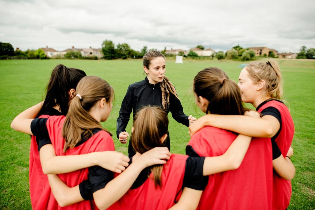 Female AIPT sports coach in a black jacket speaking to a huddle of female athletes wearing red bibs on an outdoor grass sports field.