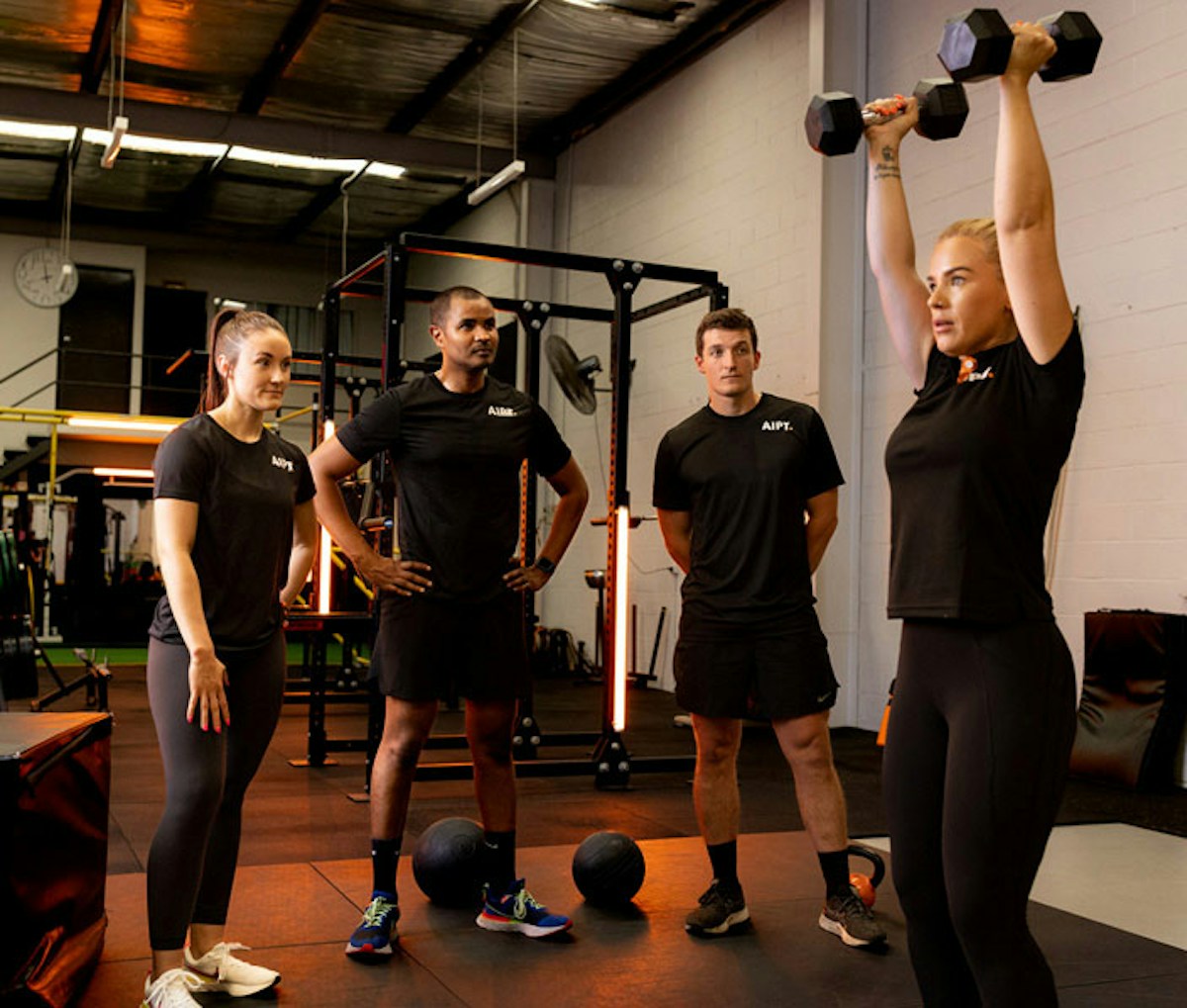 Three AIPT fitness students in black branded shirts standing and watching a blonde female student lift two black dumbbells overhead in a commercial gym setting.