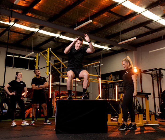 A male AIPT fitness student in a black branded shirt performs a box jump onto a large black plyo box in a commercial gym setting, while a female student and two other peers observe.