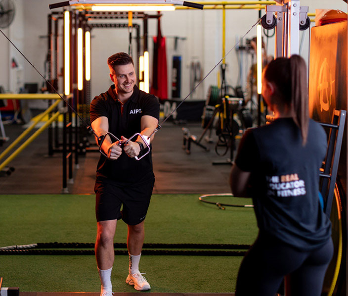 Male AIPT fitness student in a black branded polo shirt using cable crossover equipment in a gym, while a female peer in a matching shirt observes from the foreground.