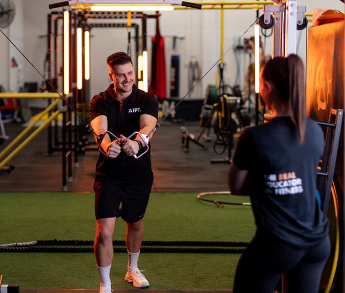 Male AIPT fitness student in a black branded polo shirt using cable crossover equipment in a gym, while a female peer in a matching shirt observes from the foreground.
