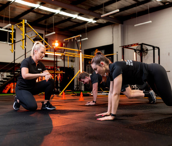 Female AIPT instructor crouches on the gym floor timing students while a female student performs a plank in the foreground during a practical fitness education session.