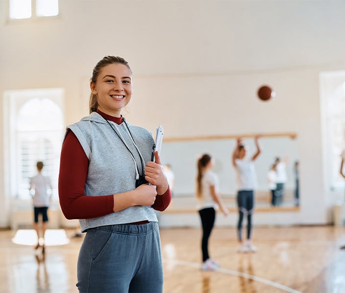 Smiling female sports coach holding a clipboard and whistle in an indoor sports hall during an Australian Institute of Personal Trainers (AIPT) training session, with other students practicing in the background.
