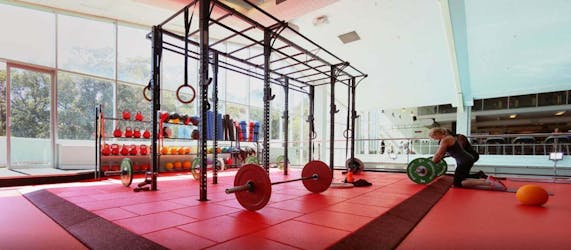 Interior view of a Fitness First gym training partner facility featuring a red and black functional turf flooring area, barbells, a large rig, and a student working out in the background.