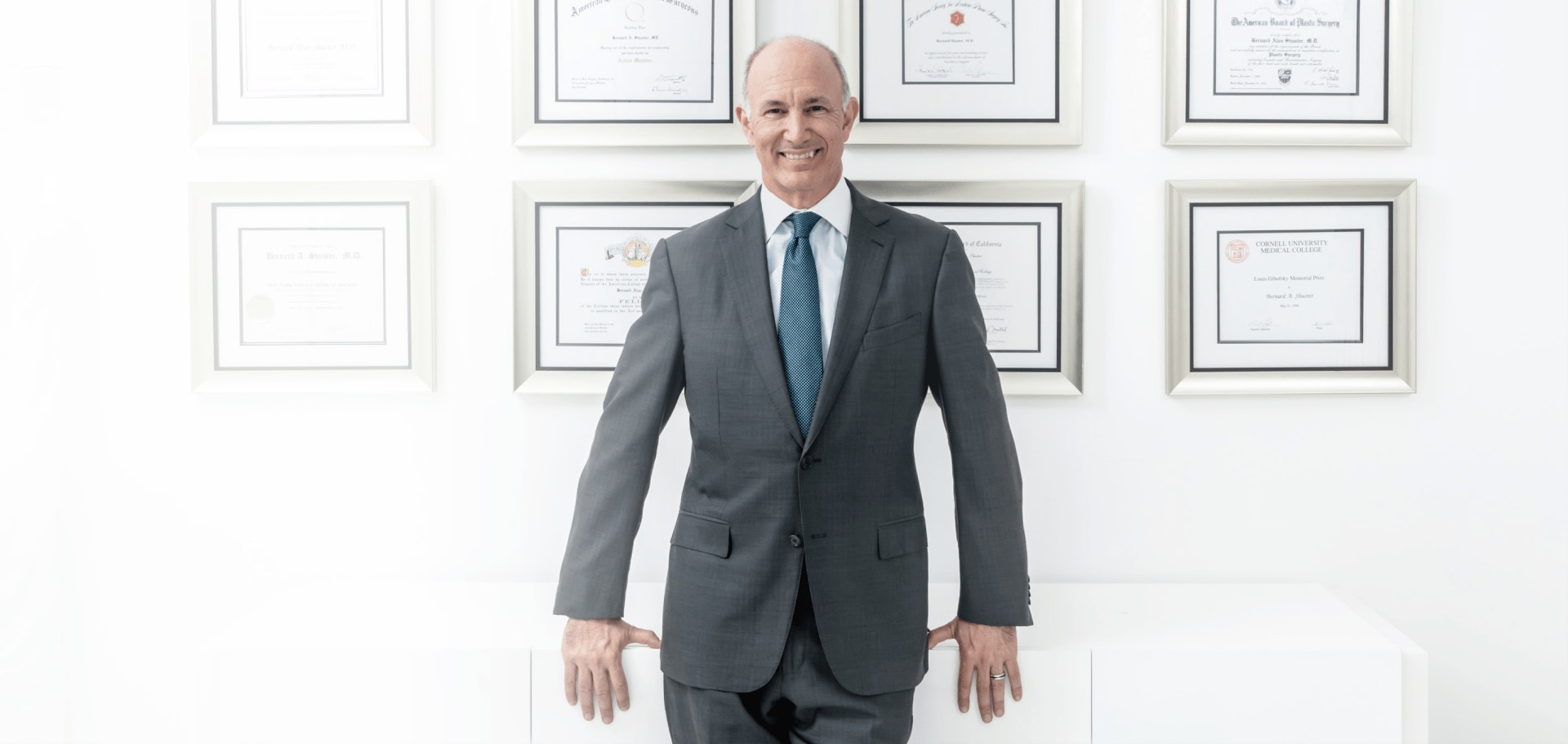 Dr. Shuster standing in his office smiling with his certificates on the wall behind him.