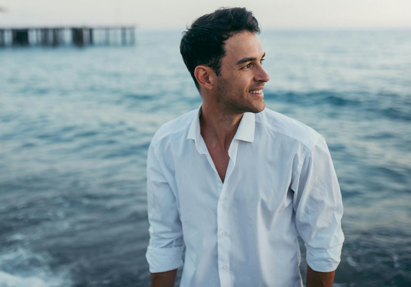 man with dark hair wearing a white button down shirt by the ocean