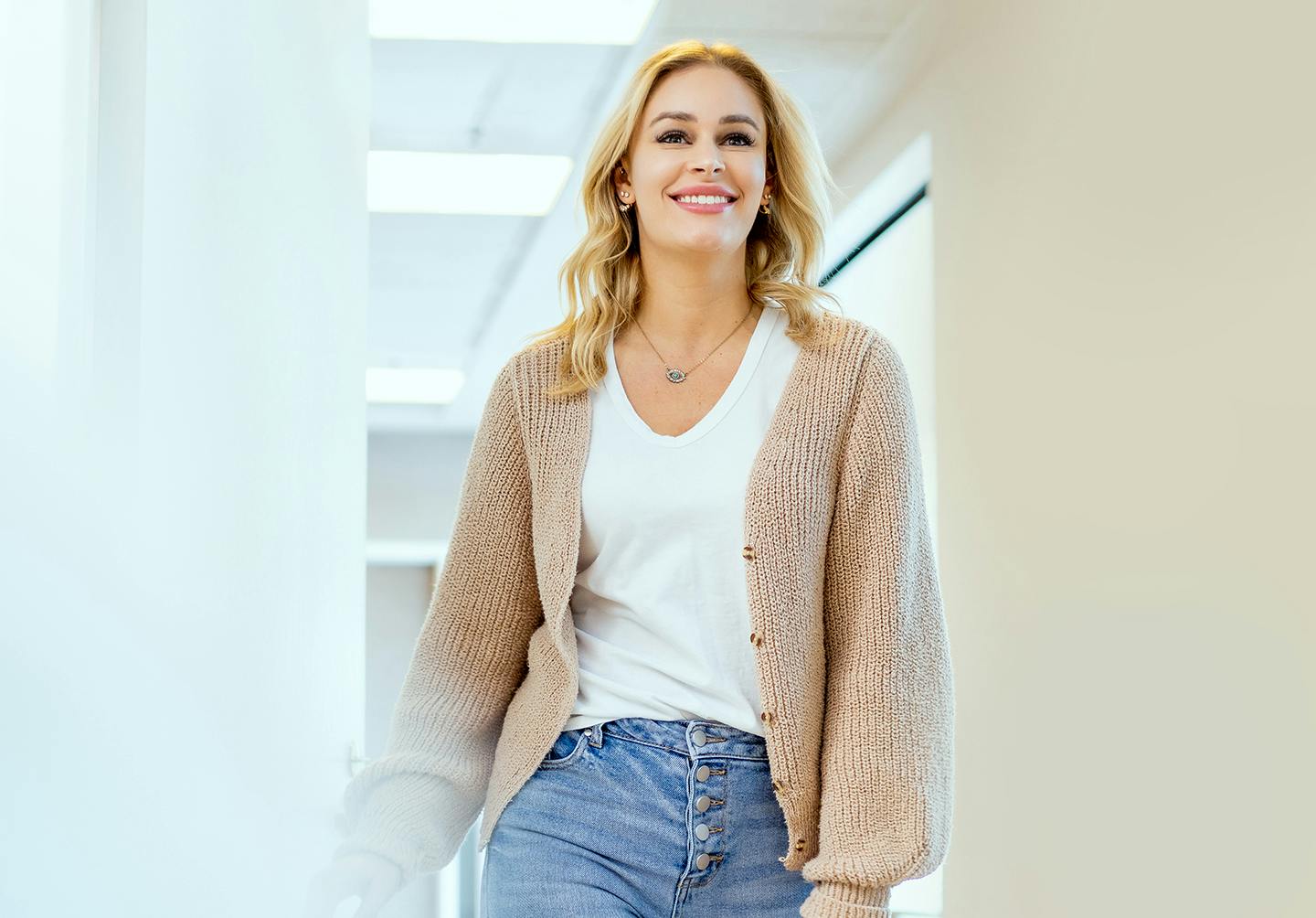 woman walking down a hallway in a white shirt, tan cardigan, and blue jeans
