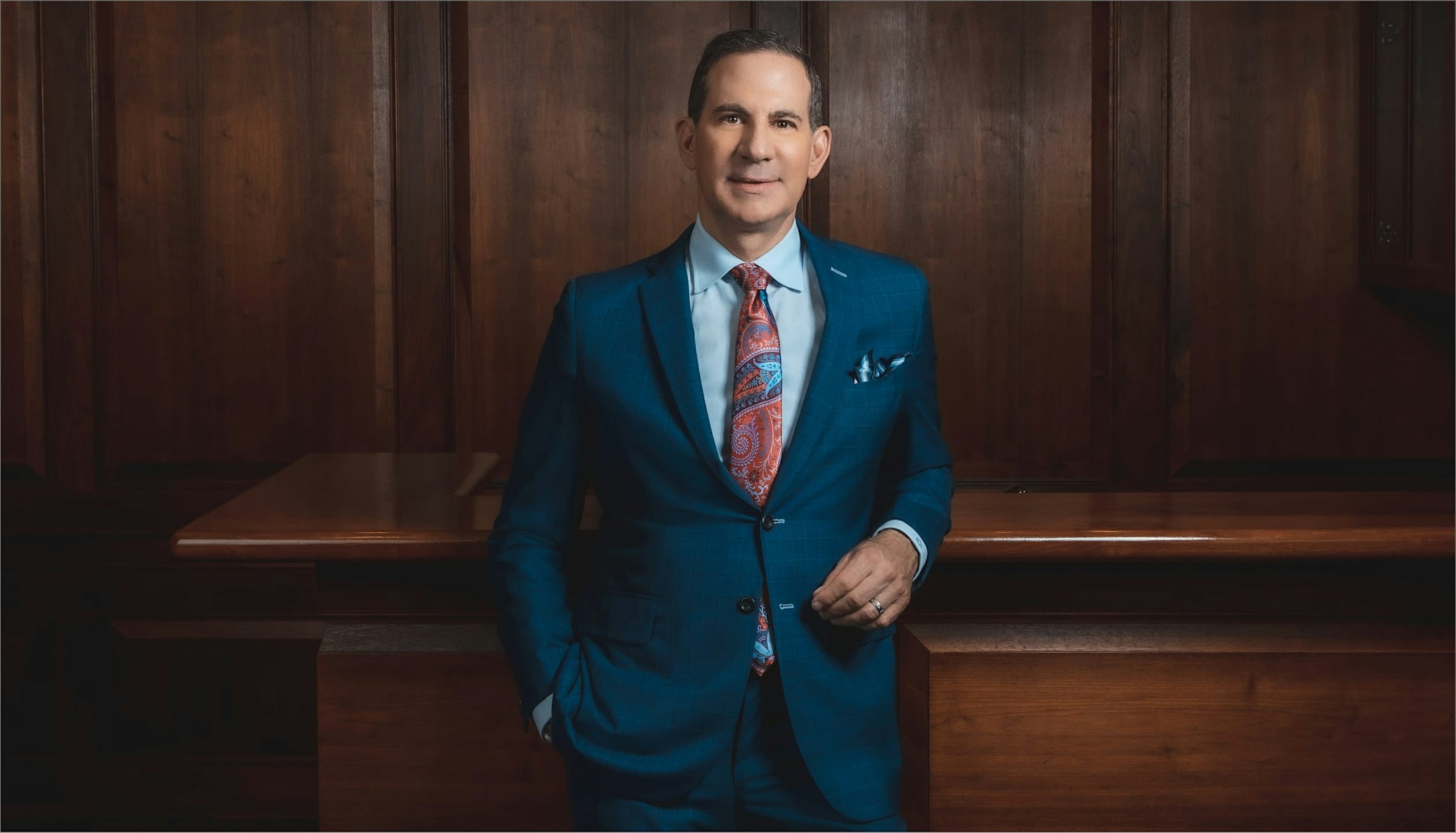 Man standing in front of a large wooden desk wearing a blue suit and looking into the camera