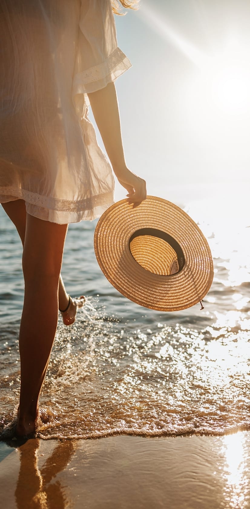 Woman standing on the beach holding a sunhat while kicking water into the air.