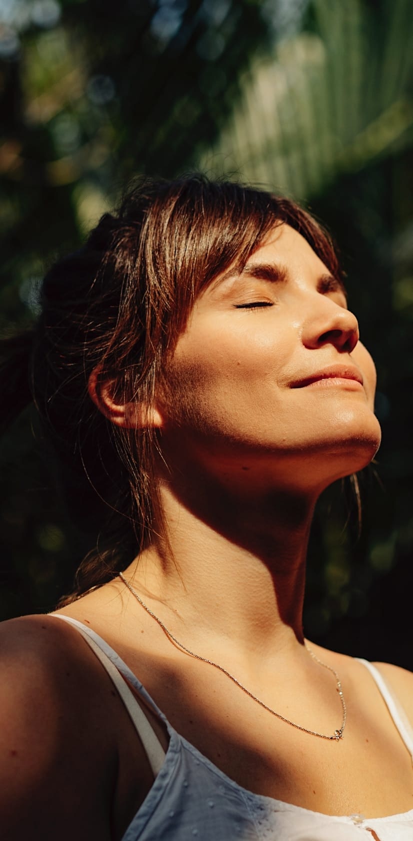 Woman with her eyes closed and looking content standing in the sun in front of a green palm leaf.