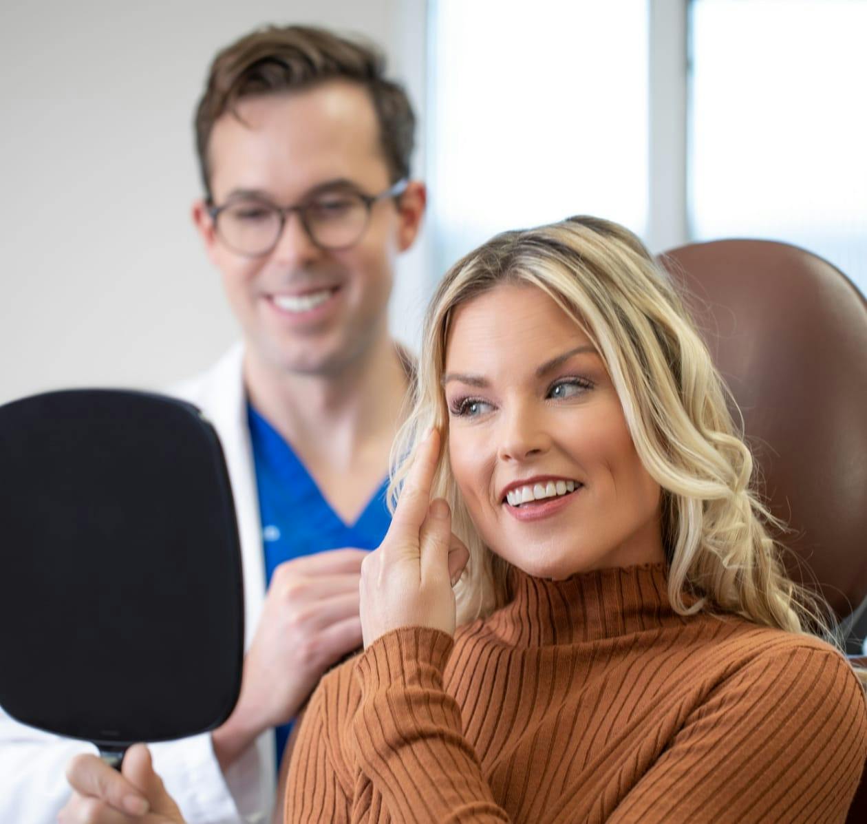 Beautiful blonde woman smiling, looking into a mirror and pointing to the side of her eye. There is a doctor behind her smiling into the mirror as well. 