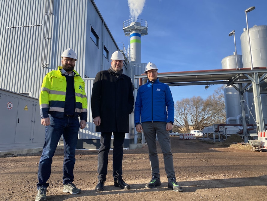 Drei Personen stehen auf einem Industriegelände vor einem großen, grauen Gebäude und hohen Tanks, alle tragen Schutzhelme, während im Hintergrund Schornsteine Rauch in den blauen Himmel abgeben.