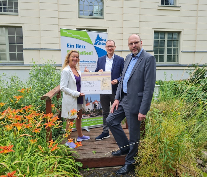 Spendenübergabe „ein Haus für Viele(s)“ Auf einem kleinen Holzsteg in einem Garten mit bunten Lilien stehen drei Personen mit einem großen Scheck vor einem Werbebanner.