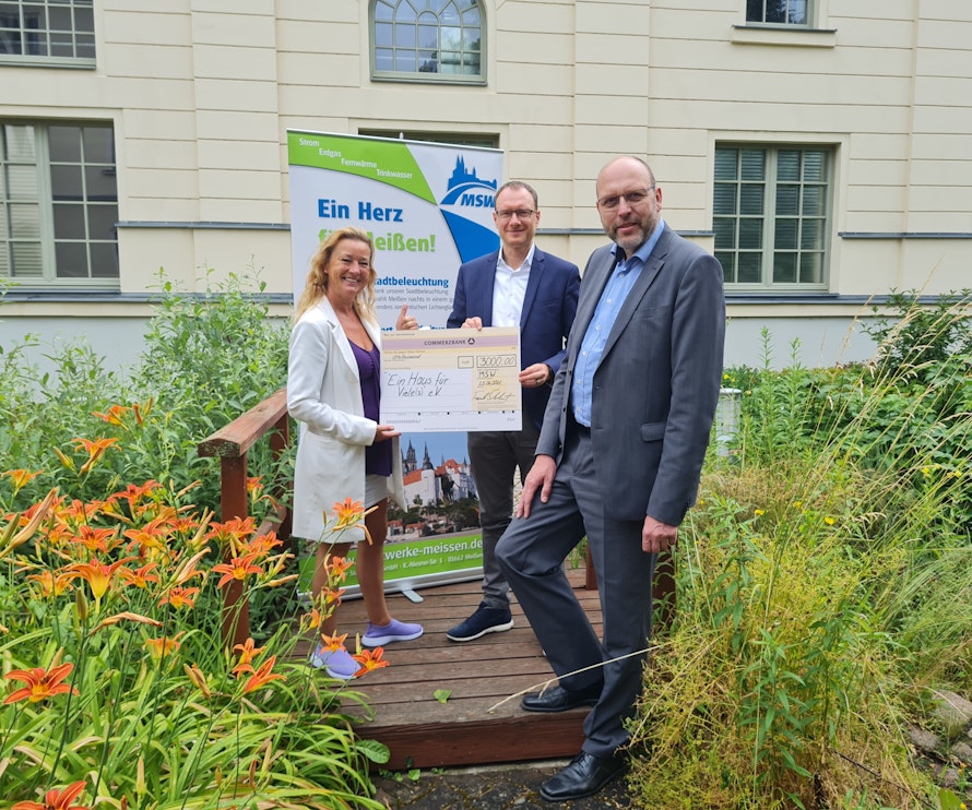 Auf einem kleinen Holzsteg in einem Garten mit bunten Lilien stehen drei Personen mit einem großen Scheck vor einem Werbebanner.