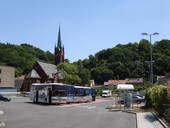 Eine Bushaltestelle in einer kleinen Stadt vor einem bewaldeten Hügel, mit einem Bus im Vordergrund und einer gotischen Kirche mit hohem Turm im Hintergrund bei klarem Himmel.