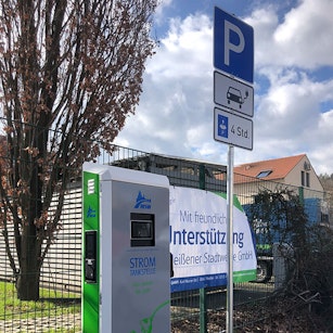Eine Stromtankstelle steht neben einem Parkplatzschild unter einem bewölkten Himmel, flankiert von einem grünen Zaun und kahlen Bäumen.