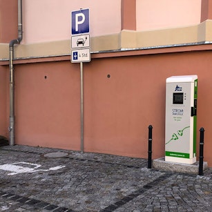 Ein Elektroauto-Ladegerät steht auf einem Kopfsteinpflasterparkplatz, markiert durch ein blaues „P“-Schild, vor einer rosa Wand, das sicher und zugänglich für Elektrofahrzeuge ist.