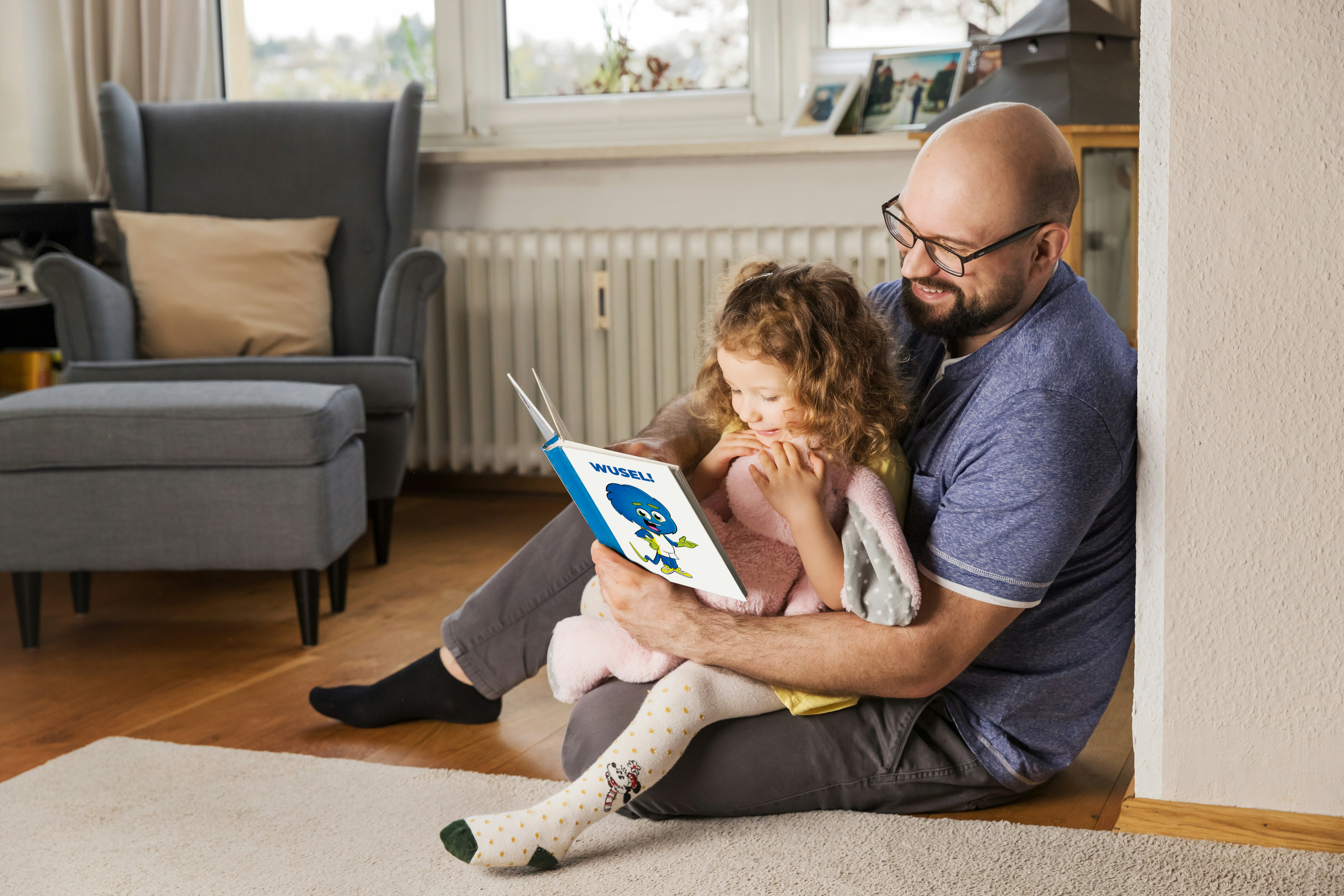 Vater und Tochter, sitzen lachend auf dem Boden und lesen in einem Kinderbuch vor einer Heizung