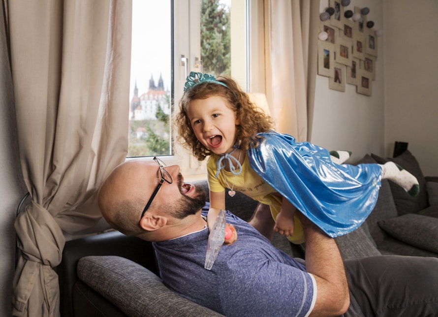 Ein fröhliches Kind in einem blauen Umhang spielt mit einem lachenden Mann auf einem Sofa, vor einem Fenster mit Blick auf ein Schloss im Hintergrund.