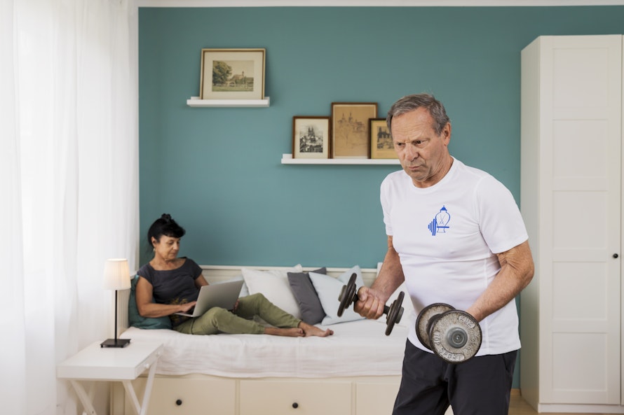 Ein älterer Mann trainiert mit Hanteln in einem modern eingerichteten Schlafzimmer mit blauen Wänden, während eine Frau entspannt auf dem Bett sitzt und einen Laptop benutzt.