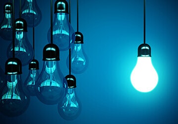Eine Gruppe von nicht leuchtenden Glühbirnen hängt neben einer einzigen eingeschalteten Glühbirne, die ein helles, blaues Licht auf einem dunkelblauen Hintergrund ausstrahlt.