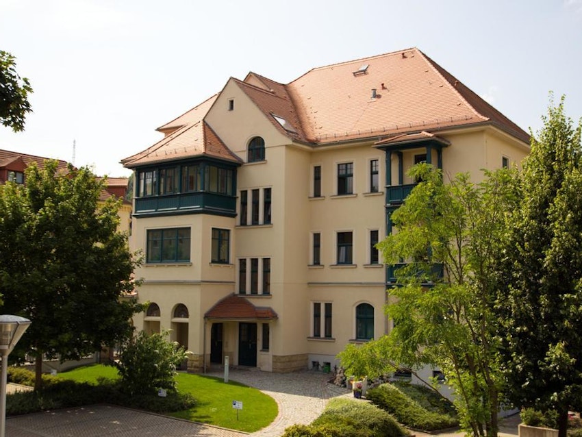 Charmantes, vierstöckiges Gebäude mit erdfarbenem Putz und roten Dachziegeln, umgeben von üppigen grünen Bäumen und einem gepflegten Gartenweg, unter blauem Himmel, der eine friedliche und einladende Atmosphäre schafft.