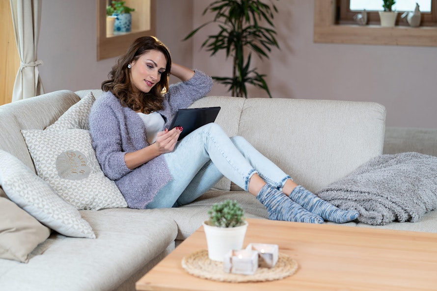 Eine Frau in lässiger Kleidung sitzt entspannt auf einem cremefarbenen Sofa, liest ein Tablet und trägt gemütliche Socken; der Raum ist hell und einladend mit Pflanzen und Kissen dekoriert.