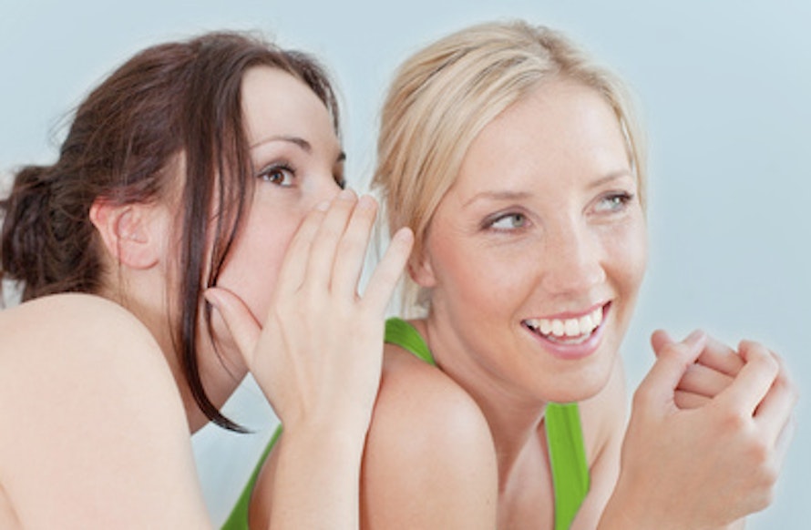 Zwei lachende Frauen in sportlicher Kleidung flüstern sich auf einem blauen Hintergrund Geheimnisse zu, strahlen Freude und Vertrautheit aus.
