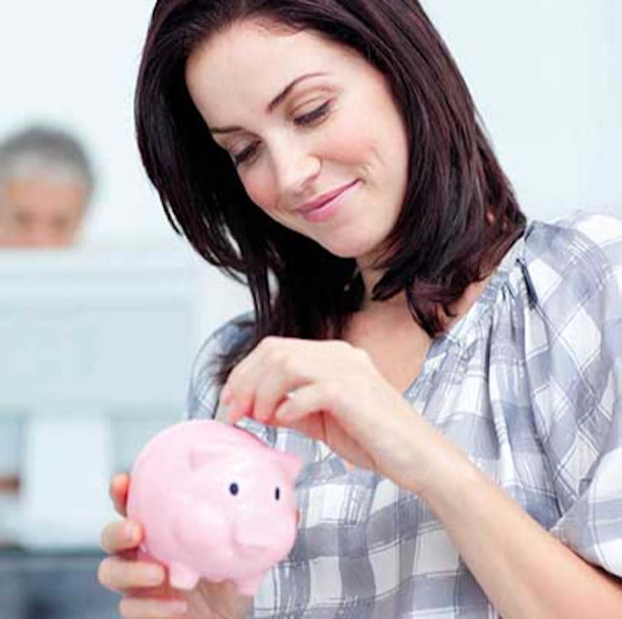 Eine Frau in kariertem Hemd steckt eine Münze in ein rosa Sparschwein, während im Hintergrund eine verschwommene Person am Computer arbeitet.