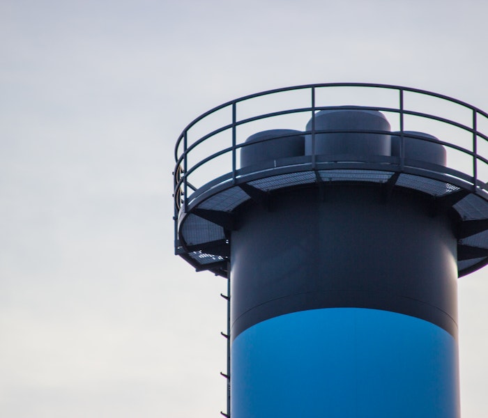 Hochgezogener Ausschnitt eines industriellen Turms mit rundem Geländer oben und blau-schwarzer Farbgebung, vor bewölktem Himmel.