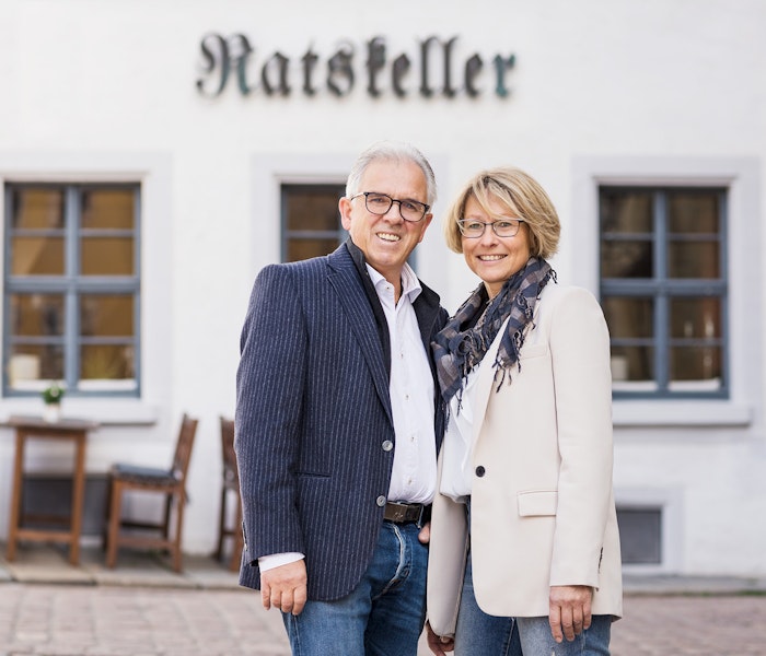 Ein lächelndes Paar steht vor einem historischen Gebäude mit der Aufschrift "Ratskeller", während sich im Hintergrund ein Tisch und Stühle im Freien befinden.