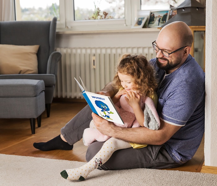 Ein Mann mit Brille und Bart liest einem kleinen Mädchen auf seinem Schoß ein buntes Kinderbuch in einem gemütlichen Wohnzimmer vor, während sie sich an ihn kuschelt und aufmerksam zuhört.