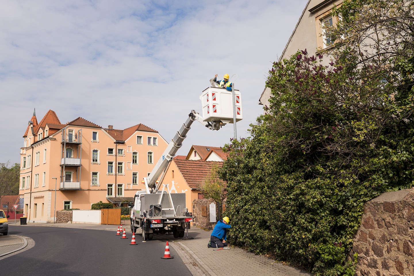 Ein Arbeiter in einem Hubsteiger repariert eine Straßenlaterne, während ein weiterer Arbeiter am Boden wartet, umgeben von Wohnhäusern und einer Hecke.