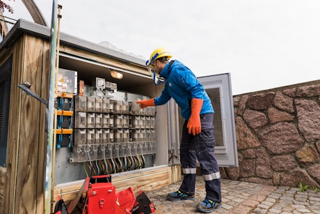 Ein Arbeiter in Schutzkleidung und Helm überprüft eine geöffnete elektrische Schalttafel im Freien, während er orangefarbene Sicherheits-Handschuhe trägt.
