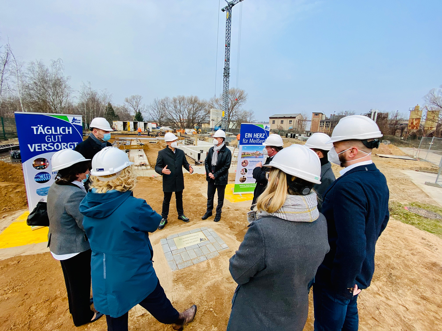 Die Gruppe von Menschen in Schutzhelmen versammelt sich um eine Baustelle und diskutiert, während im Hintergrund Werbeschilder stehen.