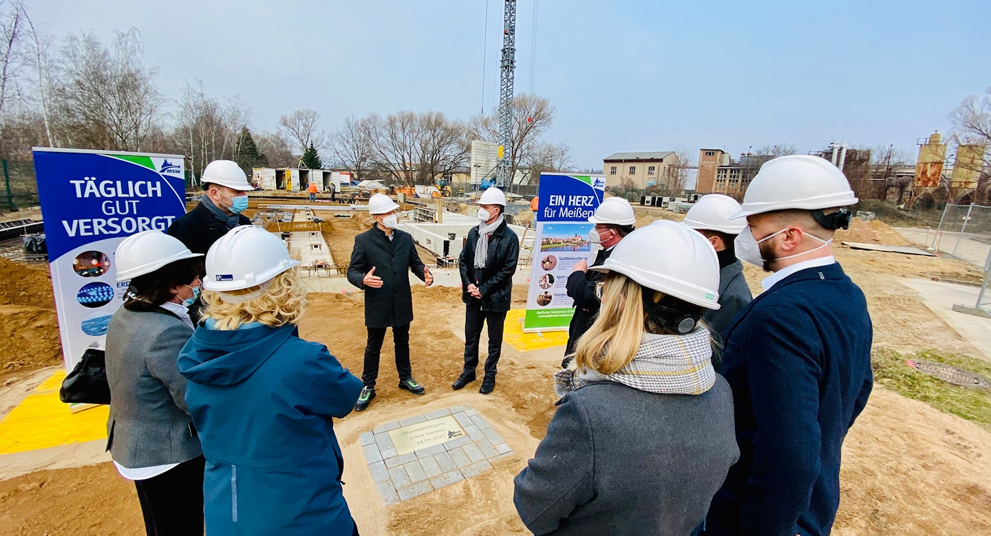 Die Gruppe von Menschen in Schutzhelmen versammelt sich um eine Baustelle und diskutiert, während im Hintergrund Werbeschilder stehen.