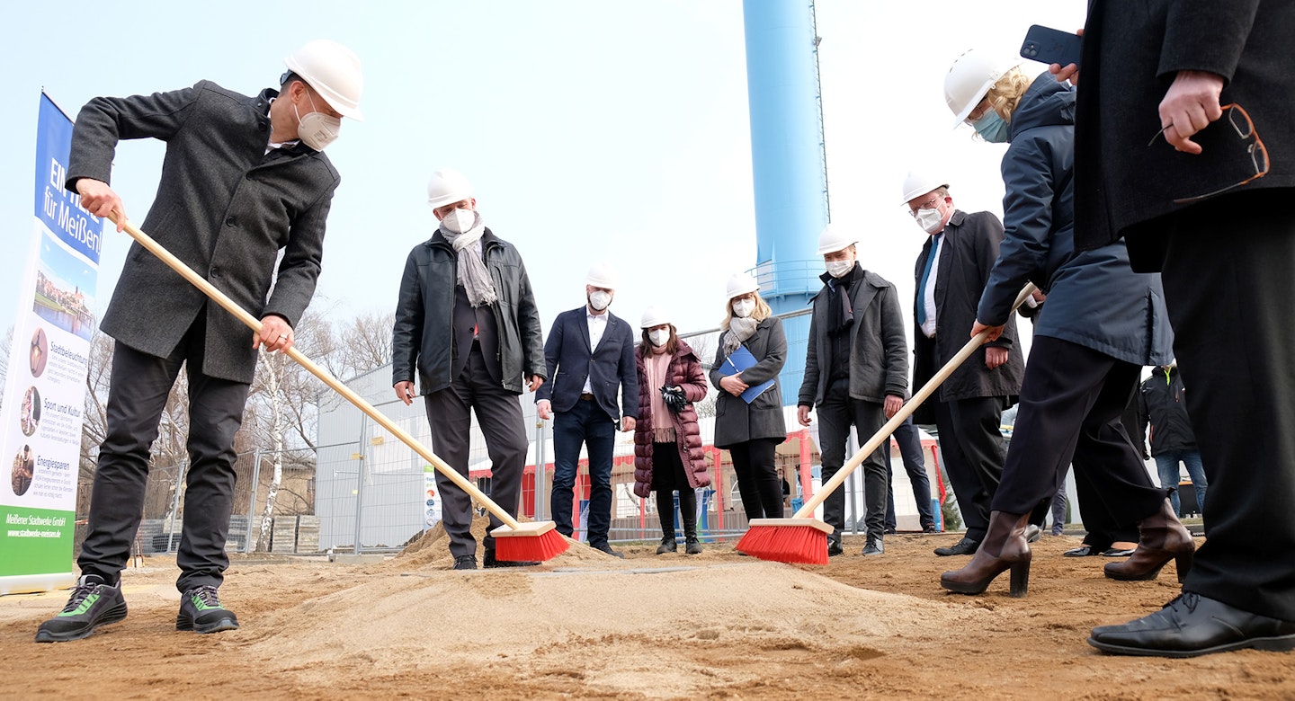 Eine Gruppe von Personen in Schutzhelmen und Gesichtsmasken nimmt an einer symbolischen Grundsteinlegung teil, wobei einige rote Besen halten, um den Sand auf einer Baustelle zu kehren.