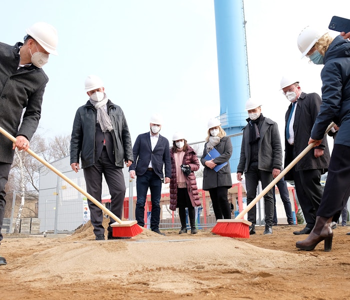 Eine Gruppe von Personen in Schutzhelmen und Gesichtsmasken nimmt an einer symbolischen Grundsteinlegung teil, wobei einige rote Besen halten, um den Sand auf einer Baustelle zu kehren.