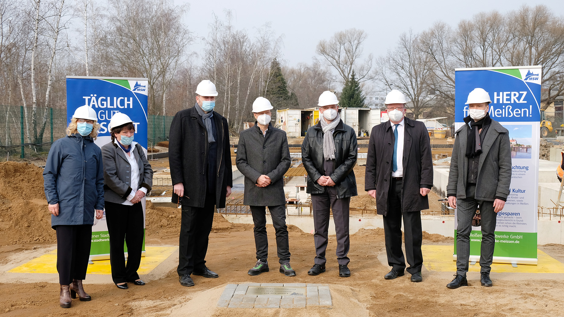 Eine Gruppe von sieben Personen in formeller Kleidung und weißen Bauhelmen steht auf einer Baustelle, flankiert von zwei bedruckten Werbebannern; im Hintergrund sind Baumaterialien und unvollendete Gebäude zu sehen.