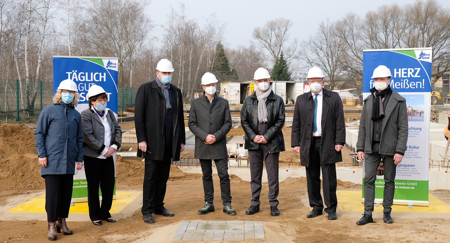 Eine Gruppe von sieben Personen in formeller Kleidung und weißen Bauhelmen steht auf einer Baustelle, flankiert von zwei bedruckten Werbebannern; im Hintergrund sind Baumaterialien und unvollendete Gebäude zu sehen.