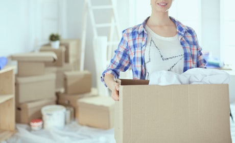 Eine Frau trägt einen Karton in einem hell beleuchteten Raum mit mehreren Pappkartons und einer Leiter im Hintergrund; es scheint ein Umzug stattzufinden.