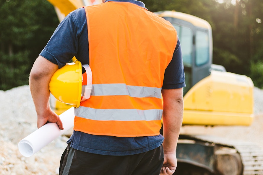 Ein Bauarbeiter in orangefarbener Warnweste hält einen Bauhelm und Baupläne vor einem gelben Bagger auf einer Baustelle.