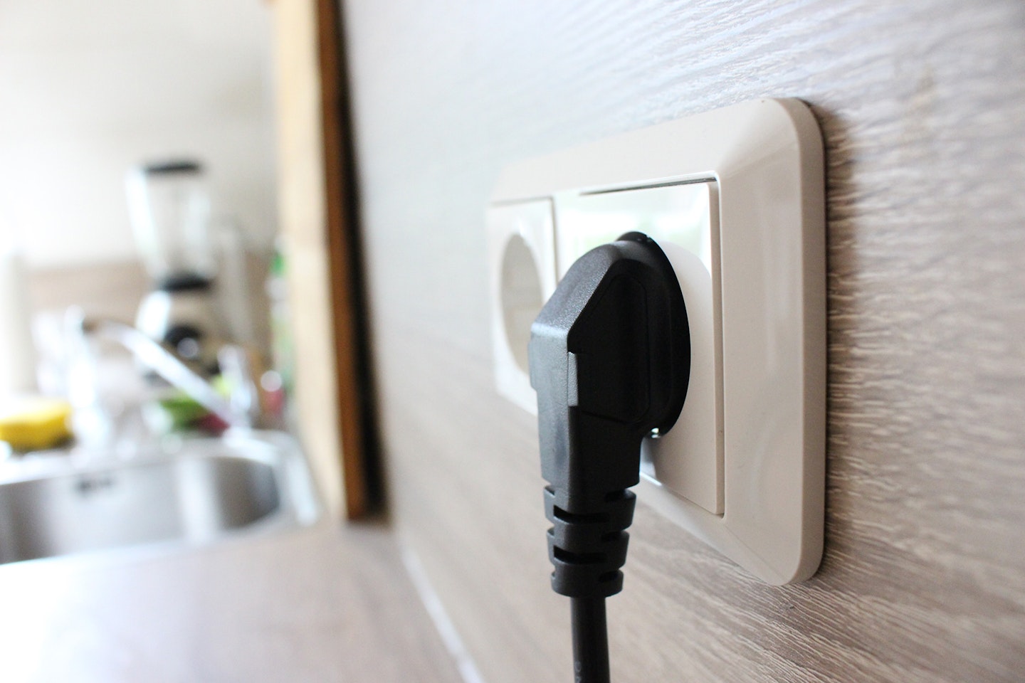 Eine Nahaufnahme eines schwarzen Steckers, der in eine weiße Wandsteckdose in einer modernen Küche eingesteckt ist, im Hintergrund ist eine verschwommene Küchenzeile zu sehen.