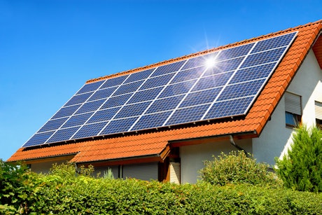 Ein modernes Einfamilienhaus mit rotem Ziegeldach ist mit einer großen Anzahl von Solarpanels bedeckt, die in der Sonne glänzen und von einem gepflegten Garten mit grünen Sträuchern umgeben sind.