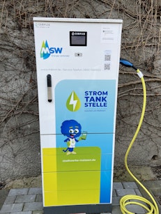 Eine Ladesäule für Elektrofahrzeuge mit dem Logo der Stadtwerke Emden und einer Karikatur eines blauen Charakters vor einer mit Efeu bewachsenen Wand, mit gelbem Ladekabel angeschlossen und bereit für die Nutzung.