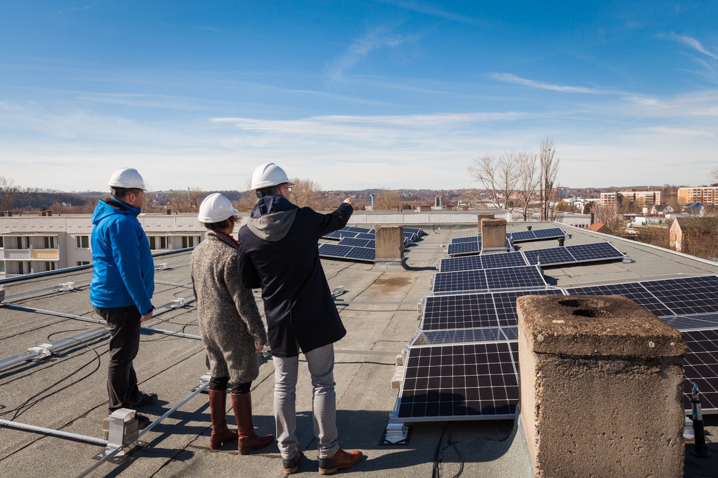 Drei Personen mit weißen Schutzhelmen inspizieren eine große Solaranlage auf einem Flachdach, während ein klarer Himmel den Hintergrund bildet.