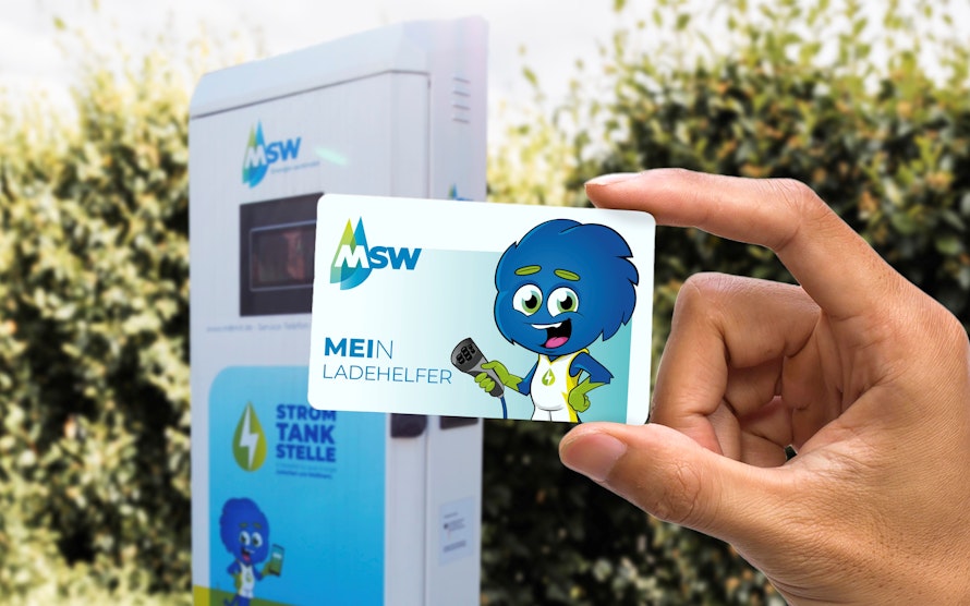 Eine Person hält eine MSW-Karte mit einem blauen Maskottchen darauf vor einer Ladestation für Elektrofahrzeuge hoch, während im Hintergrund grüne Büsche zu sehen sind.