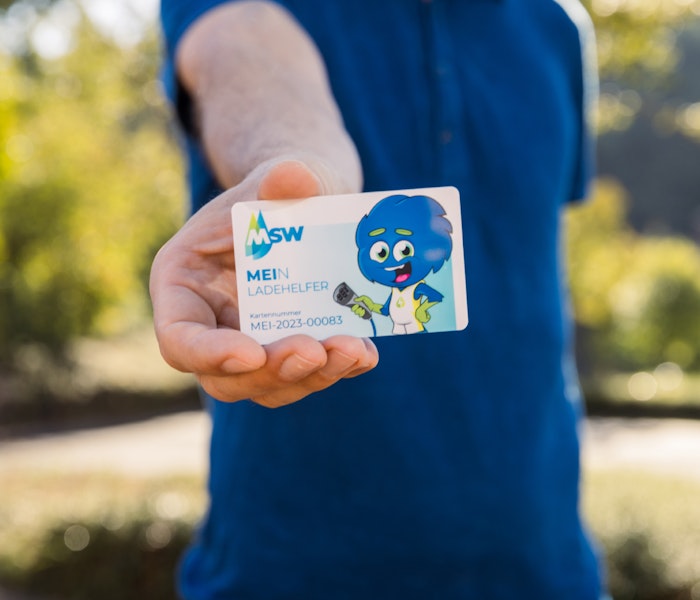Eine Person in einem blauen Hemd hält eine MSW-Einzelhandelskarte mit einem Cartoon-Charakter signifikant in die Kamera, während sie sich in einem sonnigen Park befindet.