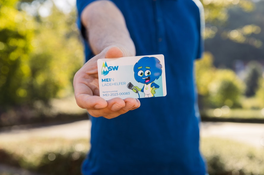 Eine Person in einem blauen Hemd hält eine MSW-Einzelhandelskarte mit einem Cartoon-Charakter signifikant in die Kamera, während sie sich in einem sonnigen Park befindet.