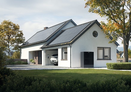 Ein modernes, weißes Einfamilienhaus mit Solarpaneelen auf dem Dach steht inmitten eines gepflegten Gartens unter einem bewölkten Himmel.